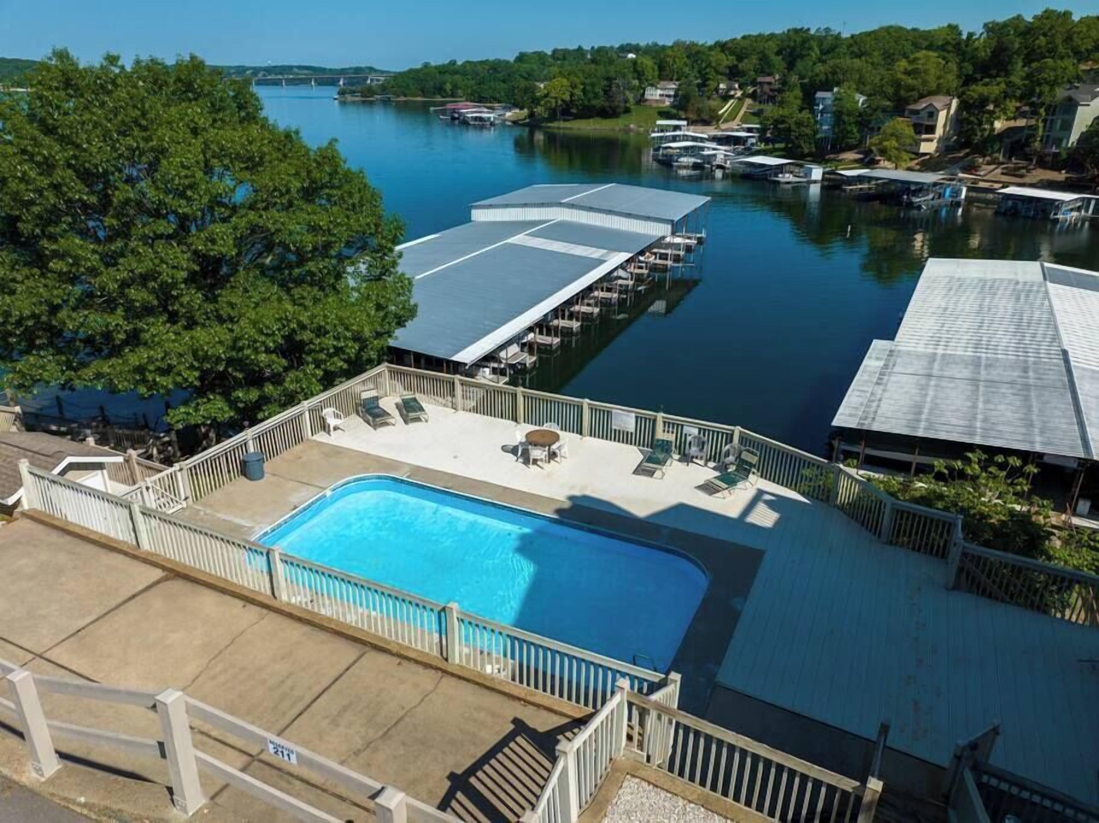 A swimming pool with a deck, surrounded by a wooden fence, and docked boats in a calm lake.