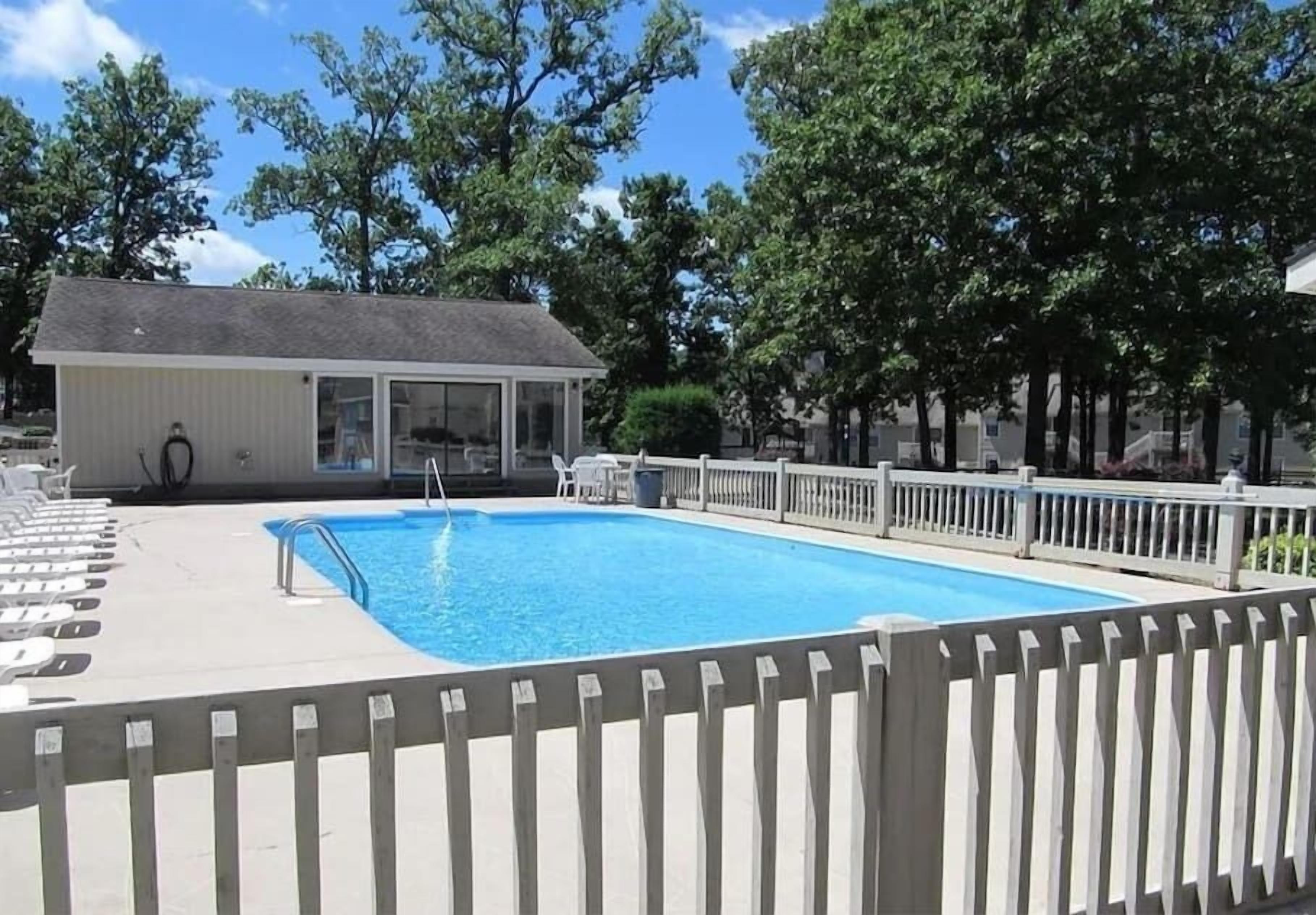 Une piscine avec une terrasse en bois, un espace salon couvert et une zone délimitée par une clôture.