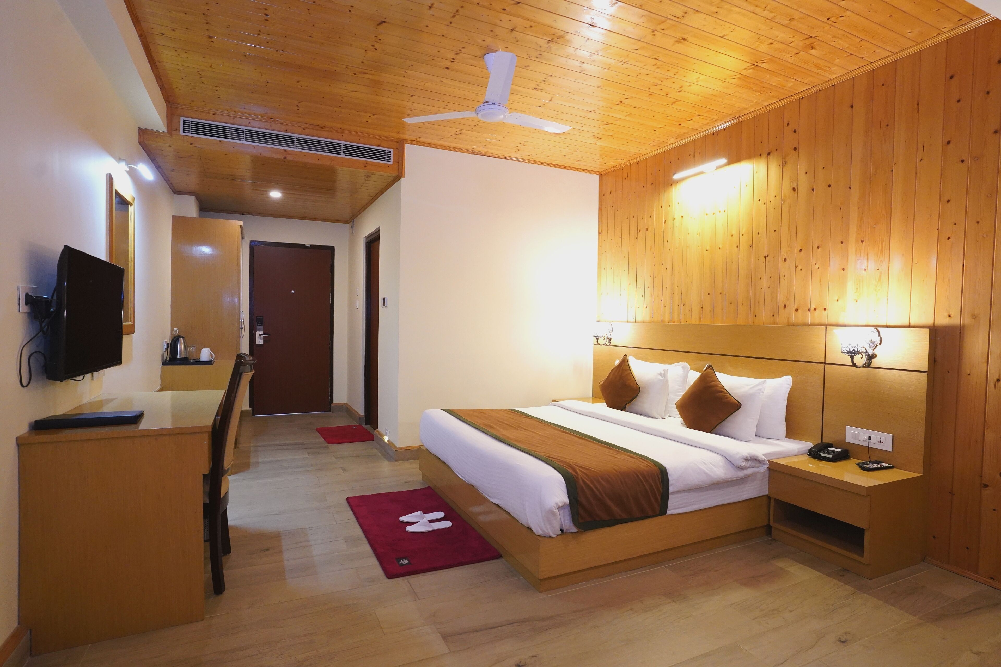 A hotel room with a wooden ceiling, a bed with white and brown bedding, a desk with a television, and a nightstand with a phone.