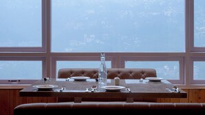 Mesa de comedor dispuesta con platos, cubiertos y una botella, frente a amplias ventanas con vista a un paisaje urbano.