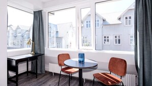 A room with a round table, two chairs, a desk, and a lamp. There are large windows with curtains, and a radiator is visible. - Svala Downtown Studio Twin beds (Reykjavík)