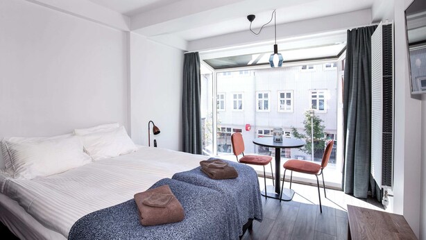 A hotel room with a large bed, two bedside tables, a small round table with chairs, and a view of buildings through the window. - Svala Downtown Studio Queen bed (Reykjavík)