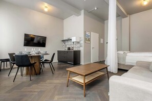 A modern living room with a sofa, a dining table with chairs, a kitchen area with cabinets, and a television mounted on the wall.