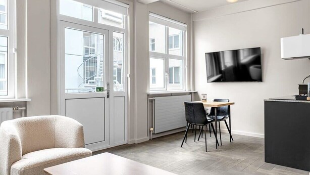 Une pièce moderne et bien éclairée, comprenant un coin repas, un téléviseur fixé au mur et une grande fenêtre laissant entrer la lumière naturelle.