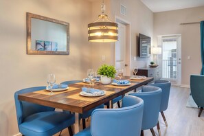 A dining area with a wooden table set for six, blue chairs, a hanging light fixture, a mirror, and a television on the wall. - VCR2-202 - 2nd Floor Unit (Orlando)