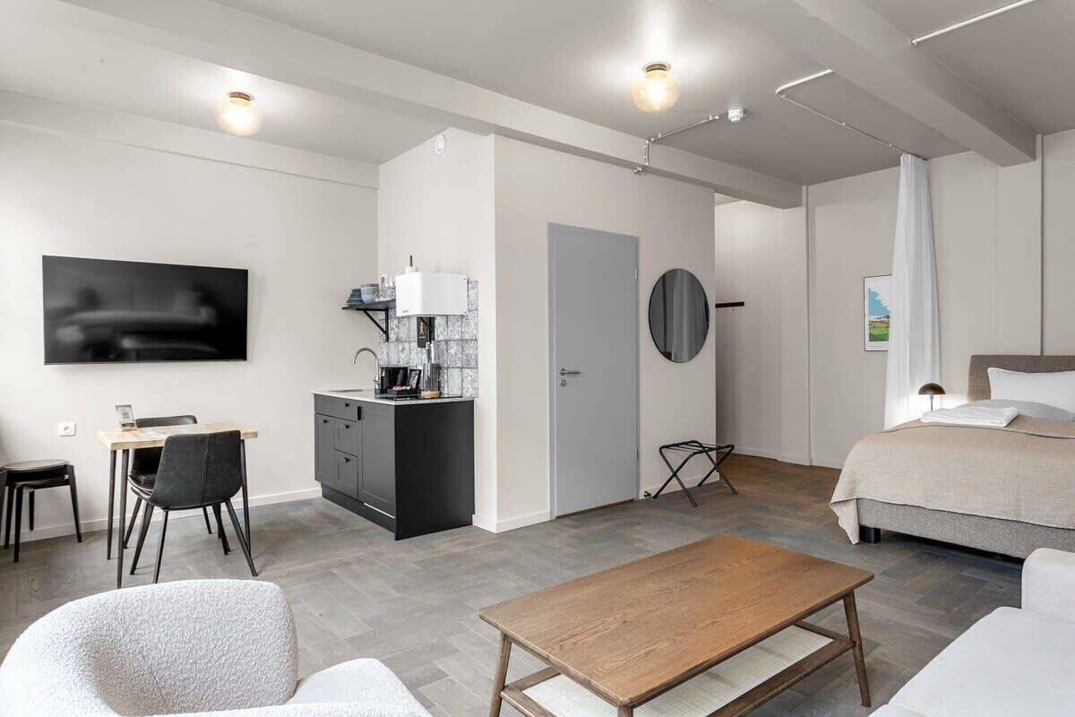 A modern hotel room with a bed, a desk with a chair, a small kitchenette, and a TV mounted on the wall.