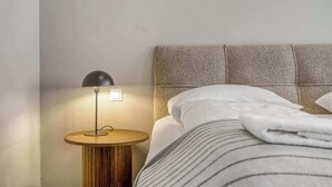 Cama bem arrumada com lençóis brancos e roupa de cama listrada, mesa de cabeceira redonda de madeira com abajur preto e cabeceira texturizada.