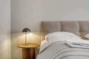 Una cama bien hecha con sábanas blancas y ropa de cama a rayas, una mesita de noche redonda de madera con una lámpara negra y un cabecero con textura.