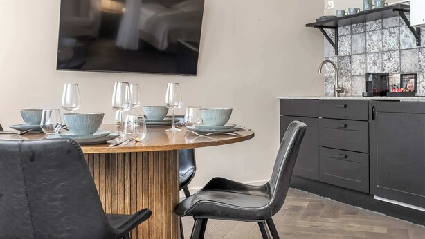 Uma sala de jantar moderna com uma mesa redonda de madeira, cadeiras pretas e uma televisão na parede.