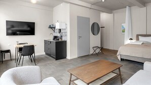 Quarto de hotel moderno com cama, escrivaninha com cadeira, pequena cozinha e TV fixada na parede.