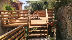 A wooden deck with a table and chairs, surrounded by a stone wall and greenery. - Appartement T2 Dans Villa à la Campagne (Solliès-Toucas)