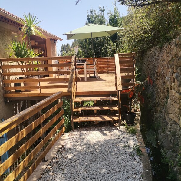 Une terrasse en bois avec une table et des chaises, entourée d’un mur de pierre et de verdure.