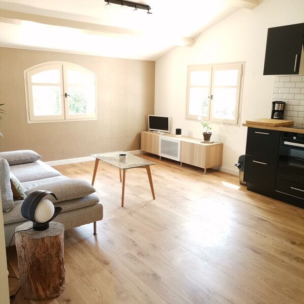 Een moderne woonkamer met een bank, een glazen salontafel en een televisie. Er is een keukenhoek met een fornuis en kasten.
