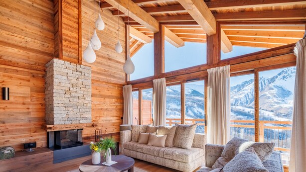 Ein gemütliches Wohnzimmer mit Kamin aus Stein, Holzbalken und großen Fenstern mit Blick auf die Berge.