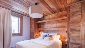 Ein Schlafzimmer mit Holzboden, einem Bett mit weißer Bettwäsche, einem Fenster mit Vorhängen und einem Nachttisch mit Lampe.
