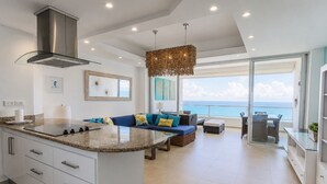A modern kitchen with a central island, a dining area with a view, and a living room with a sofa and cushions.
