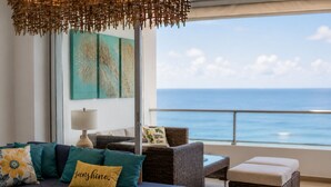 A living room with a sofa, cushions, a glass coffee table, wicker furniture, a lamp, and a view of the ocean.