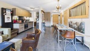 A modern kitchen with wooden cabinets, a dining area with a long table and chairs, and a living area with a sofa and a suitcase.