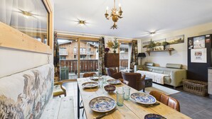 A dining area with a wooden table set for four, surrounded by chairs. A chandelier hangs from the ceiling, and there is a view of a wooden deck through a glass door.