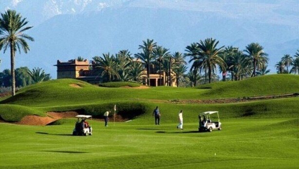 Camp de golf amb muntanyes de fons, palmeres i un edifici amb teulada plana.