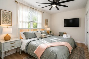 Une chambre à coucher comprenant un lit, des tables de chevet, un ventilateur de plafond, une télévision et une fenêtre avec des rideaux.