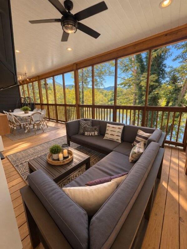 A covered deck with a sofa, coffee table, and dining area overlooking a lake.