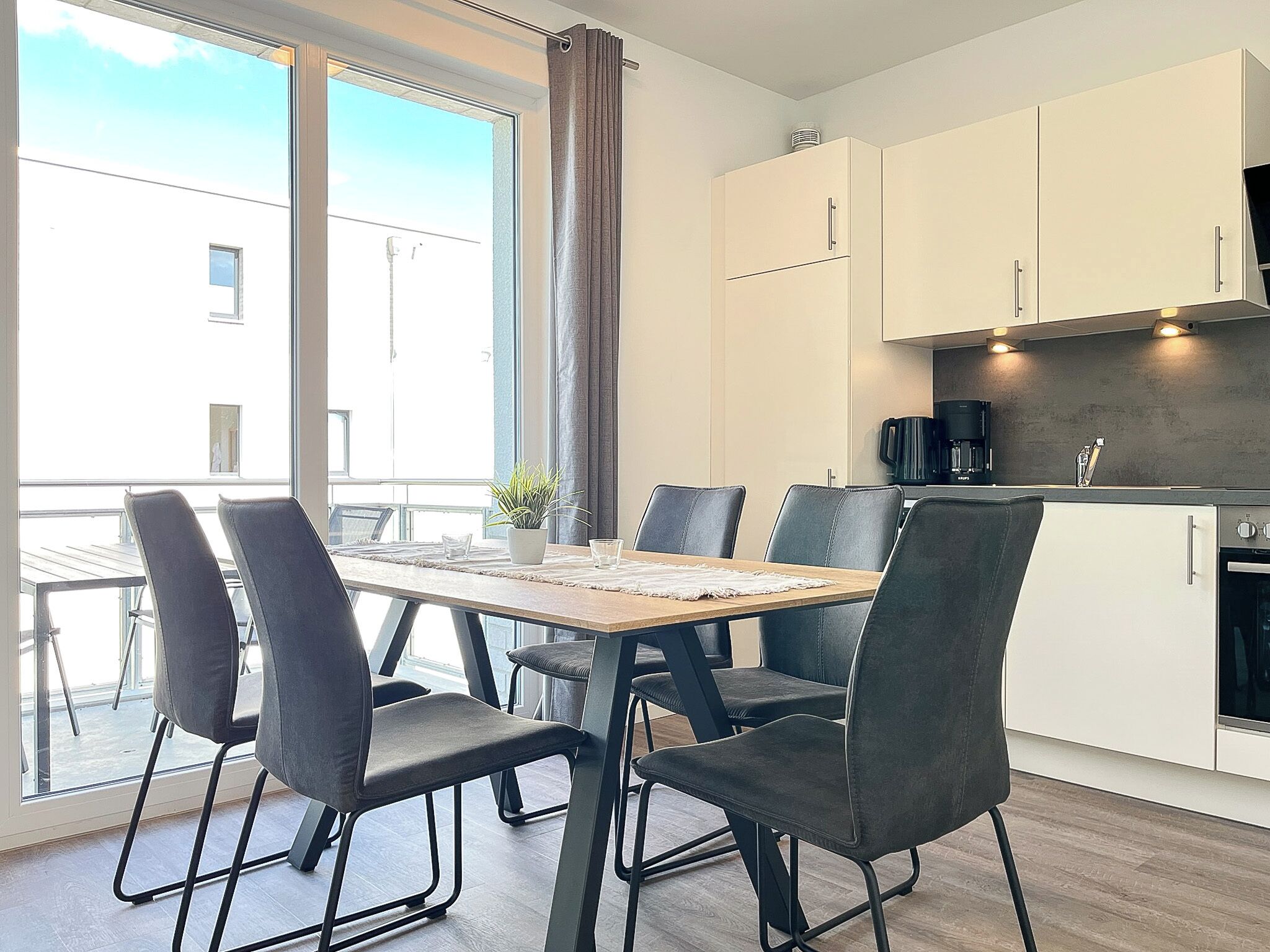 Cocina moderna con armarios blancos, una cafetera negra y un comedor con mesa de madera y sillas negras.