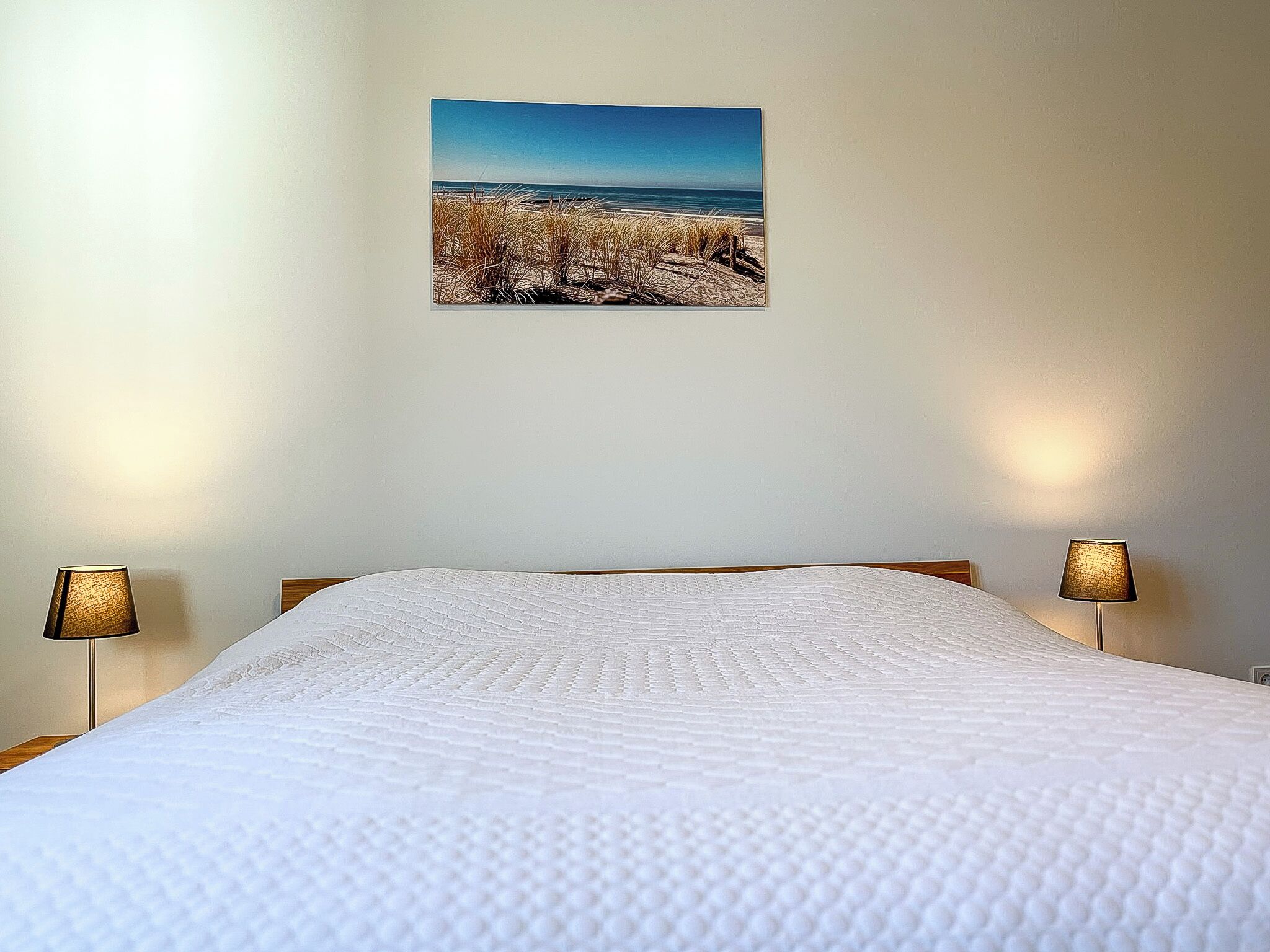 A bedroom with a bed, two bedside lamps, and a beach scene painting on the wall.