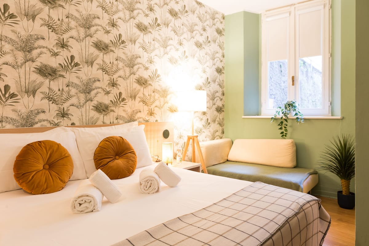 Una cama bien tendida con dos almohadas, una lámpara de noche y una ventana con una planta.
