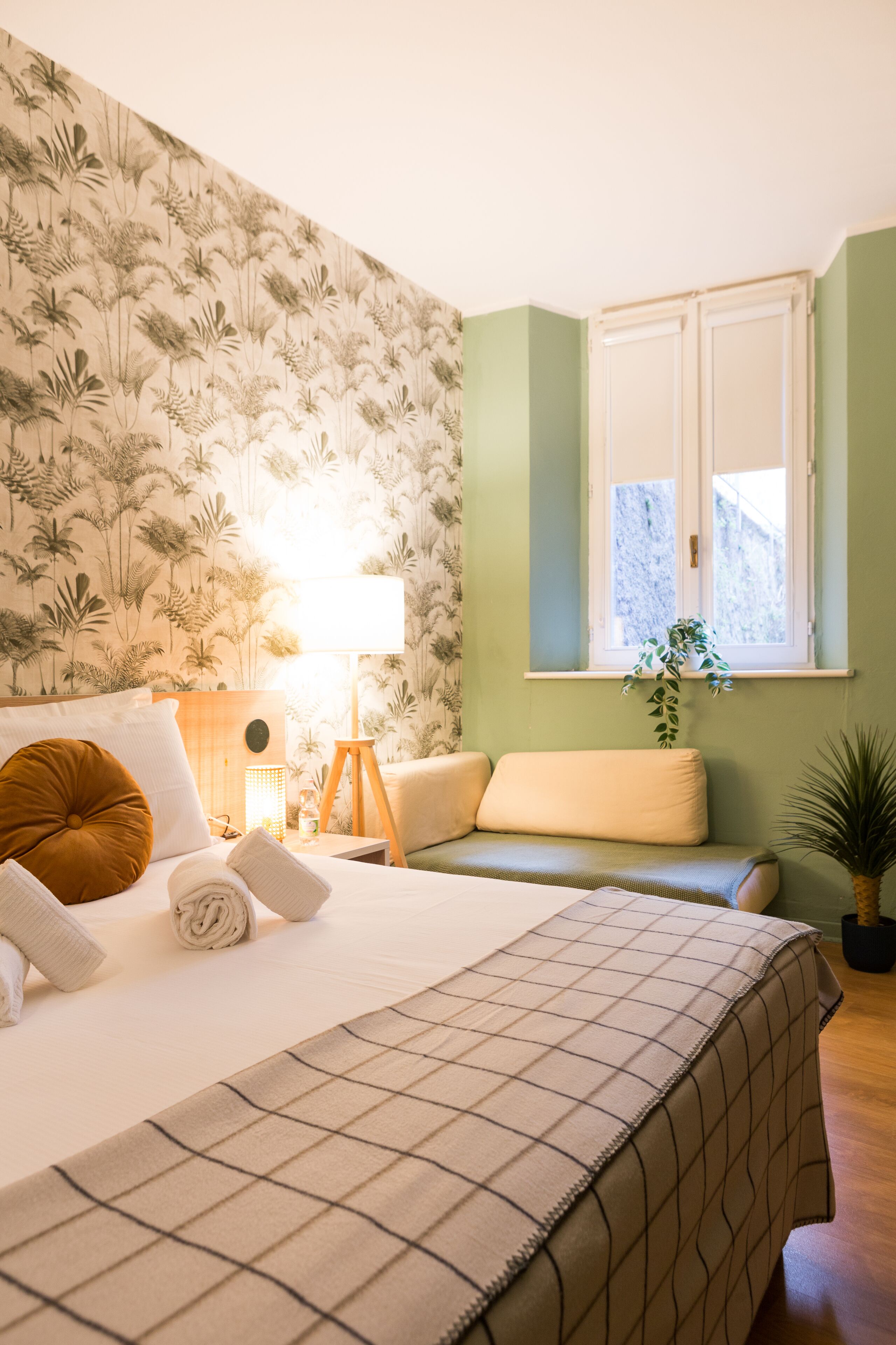 Una cama bien hecha con una manta a cuadros, una lámpara de noche y una pared verde con una ventana.