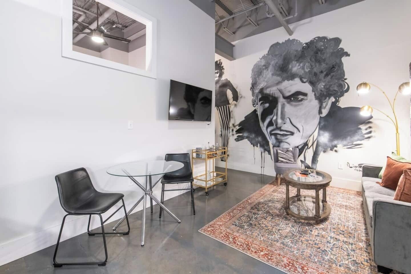 A modern interior with a round glass table, black chairs, a patterned rug, and a large abstract mural on the wall.