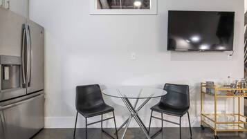 A modern kitchen with a stainless steel refrigerator, a glass table with two black chairs, a mounted flat-screen TV, and a small cart with kitchen items.