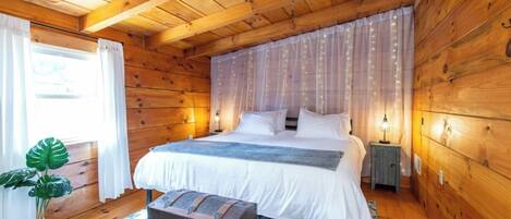 A wooden cabin bedroom with a bed, bedside lamps, a ceiling fan, and a window with curtains.