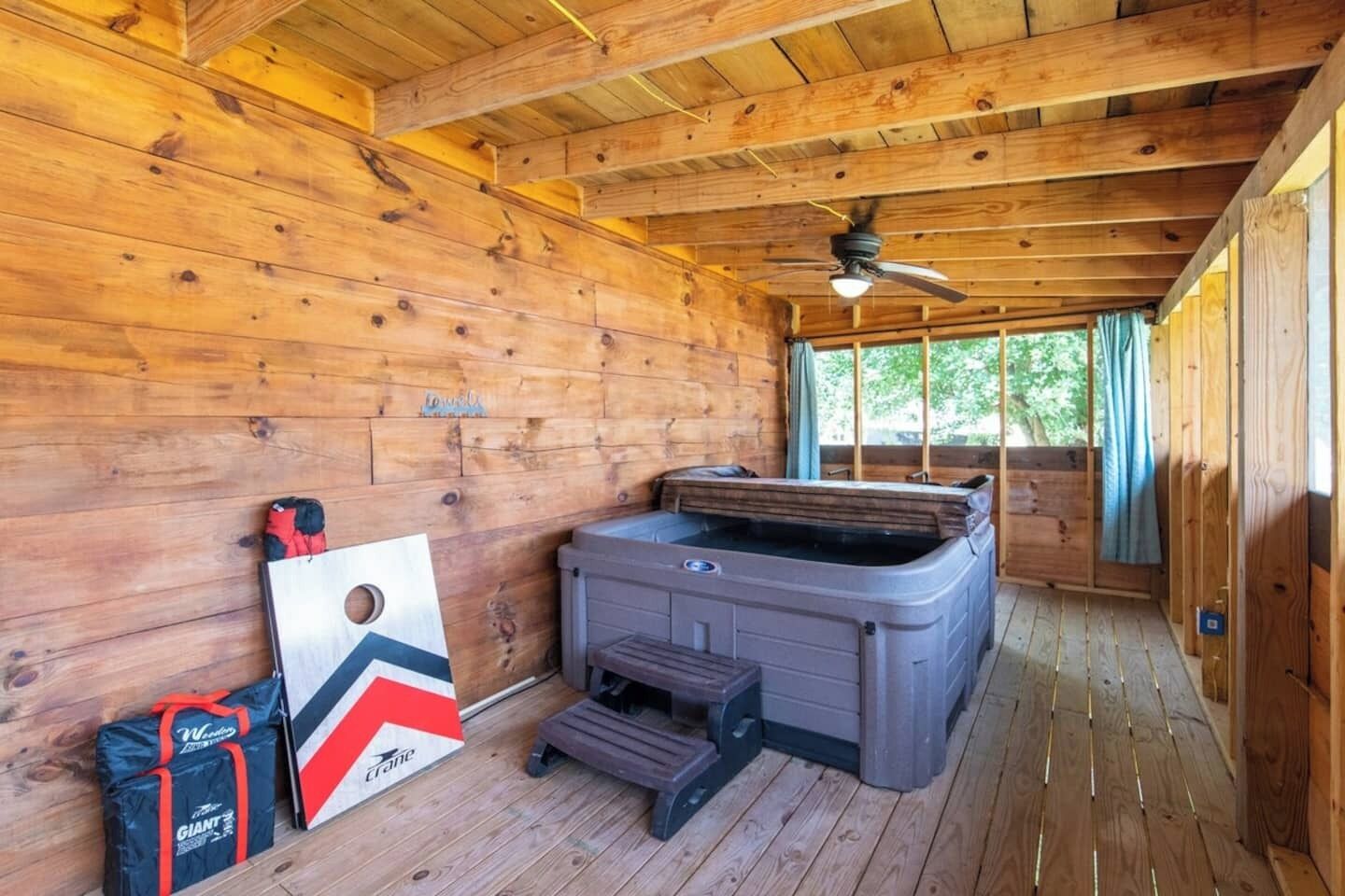 Interior de una cabaña de madera con un jacuzzi, ventilador de techo y una terraza de madera exterior.