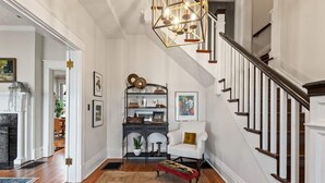 A spacious living room with a wooden floor, a fireplace, a staircase with dark wooden railings, and a chandelier.