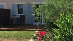 Un edificio de dos plantas con techo plano, un patio cubierto con mesa y sillas, y un pequeño jardín con flores rosadas.