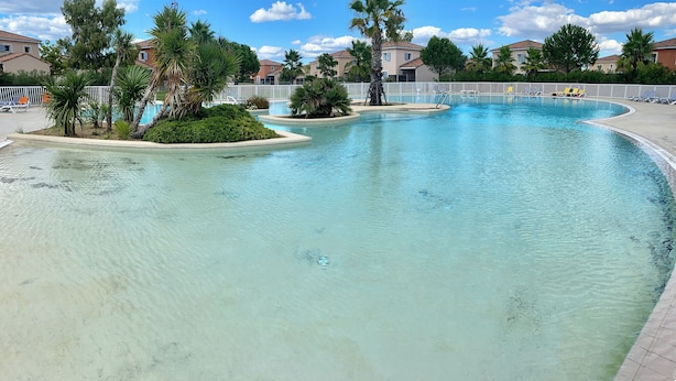 Una piscina cristalina de borde curvo, rodeada por una valla blanca, palmeras y una fila de casas al fondo.