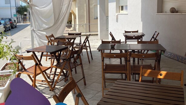 An outdoor patio area with wooden tables and chairs, surrounded by white walls and a covered section with a white canopy. - Viale Lazzaro Papi (Rimini)