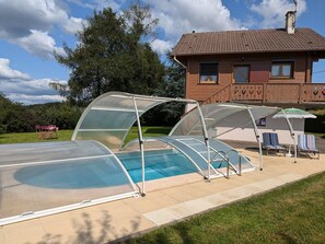 Una piscina con cerramiento de vidrio, un área de estar techada con sillones de descanso, y una casa al fondo.