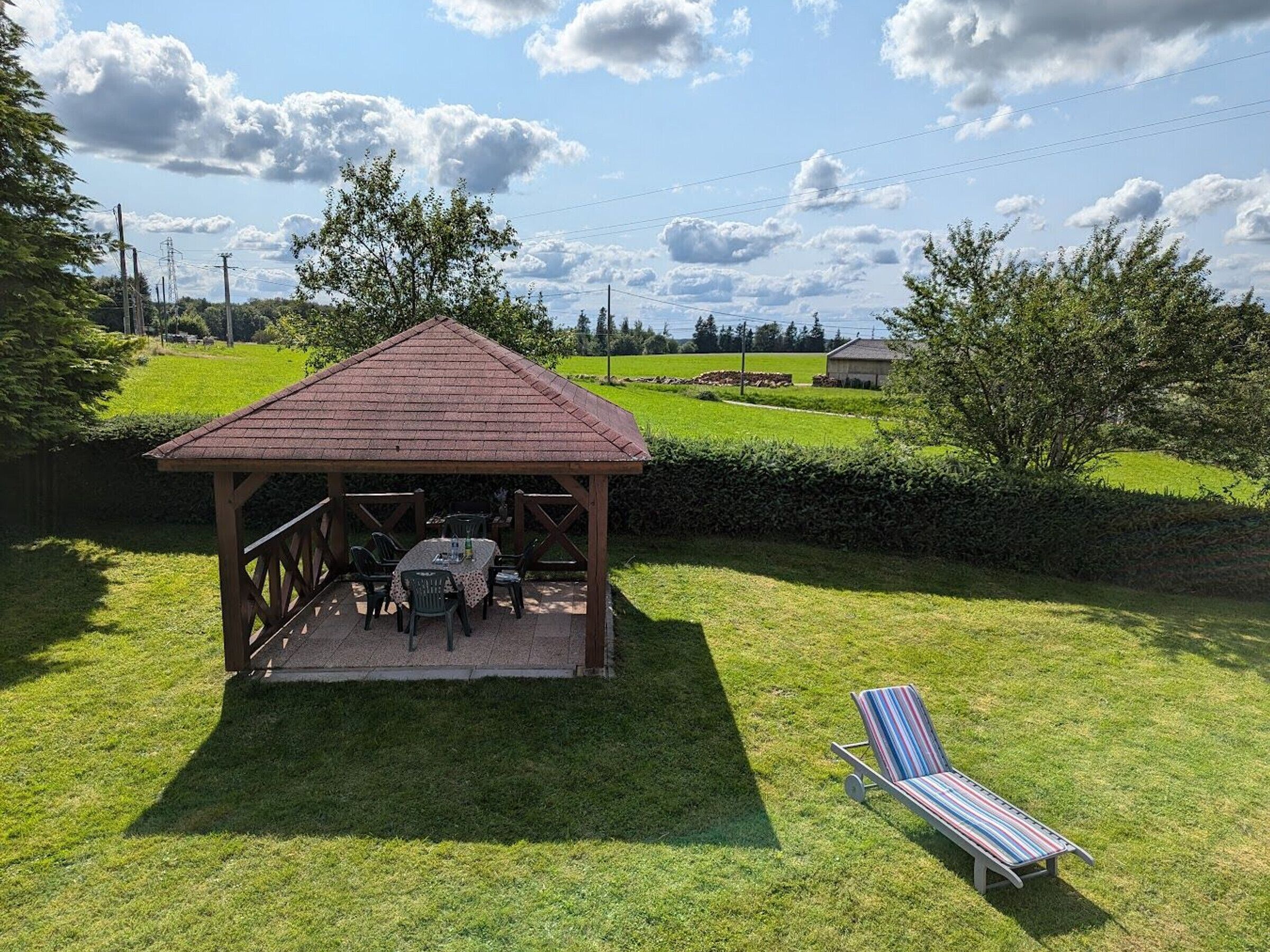 A gazebo with a table and chairs, a lounge chair, and a grassy area with trees and a clear sky.