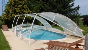En indendørs swimmingpool med gennemsigtigt tag, omgivet af en betonflisebelagt terrasse og træloungestole.