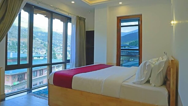 A hotel room with a large bed, a view of a cityscape through large windows, and a balcony with a railing. - The BD Hotel (Thimphu)