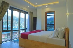 A hotel room with a large bed, a view of a cityscape through large windows, and a balcony with a railing. - The BD Hotel (Thimphu)