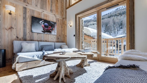 A cozy room with a wooden ceiling, a large window with a snowy mountain view, a comfortable sofa, and a rustic wooden coffee table.