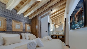 A bedroom with a large bed, wooden ceiling beams, a stone wall, and a bathroom with a mirror and sink.