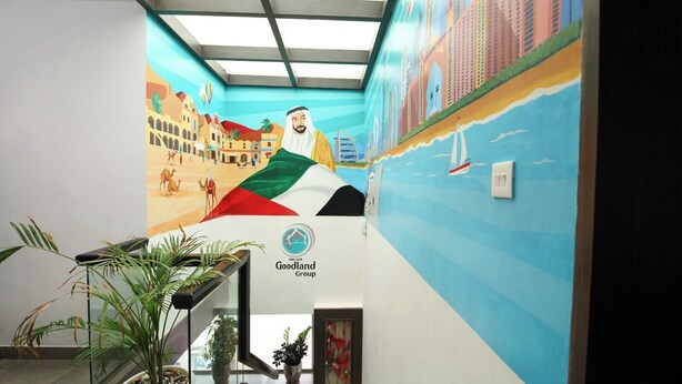 A hotel interior with a mural depicting a desert scene and a cityscape, a staircase with potted plants, and the Goodland Group logo. - Goodland Elegance (Thiruvananthapuram)