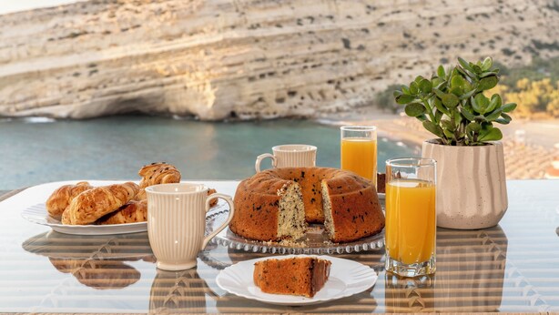 Ein Frühstückstisch mit Croissants, Kuchen und Orangensaftgläsern, vor einer malerischen Klippenlandschaft.