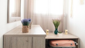 Eine Holzkommode mit einem Koffer darunter, ein Spiegel an der Wand und zwei Vasen mit Pflanzen auf einem Tisch.