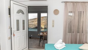 Ein Zimmer mit einem Bett, einem Tisch und Blick auf felsiges Gelände durch eine offene Tür.