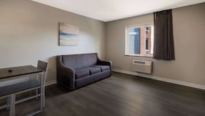 Ein modernes Wohnzimmer mit einem dunkelgrauen Sofa, einem kleinen Tisch mit einem Stuhl, einem Gemälde an der Wand und einem Fenster mit Vorhängen.